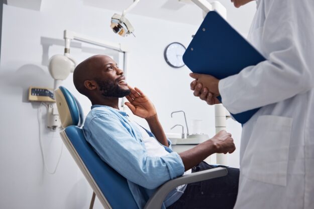 A man sitting in a dental chair gestures toward his jaw while talking to a dentist holding a blue clipboard.